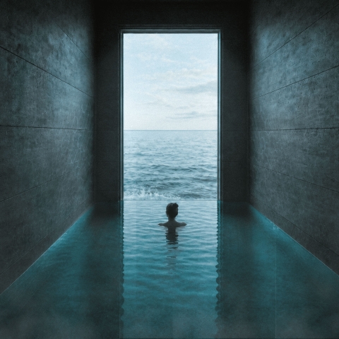 A person standing in an indoor pool framed by dark stone walls, looking out toward the bright ocean beyond.