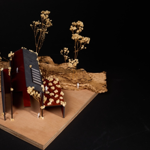 A small architectural model with angled grey and red panels set in textured brown terrain, surrounded by tiny dried plants.