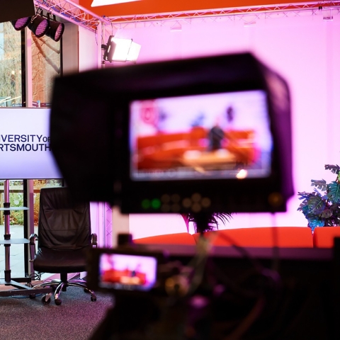 A picture of podcasting hardware in a recording studio with a screen in the background with the university of portsmouth logo displayed