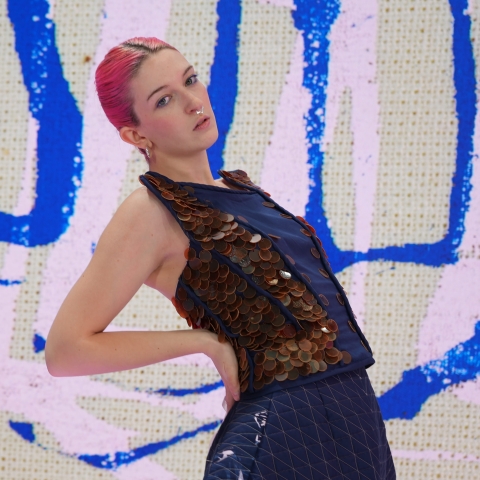 Garment by Aimee Hurst of model with pink hair doing a lean back pose. The Garment is blue and has brown chain mail style top