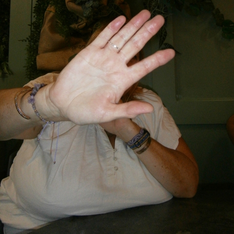 A person indoors raises an open hand toward the camera, partially blocking their face. They wear a white shirt and bracelets, with green foliage visible in the background.