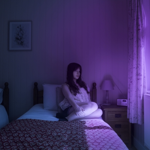 A person sits on a bed in a dimly lit wooden‑panelled room, illuminated by vivid purple and blue light from a nearby window. A patterned blanket, a nightstand with a lamp, and a framed picture complete the moody scene.
