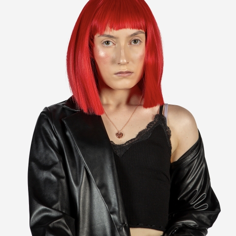 A close‑up black‑and‑white image showing part of a person with straight, bright red hair cut into a blunt bob with bangs. They wear a black leather jacket draped off one shoulder, a black lace‑trimmed camisole, and a small pendant necklace, set against a plain white background.