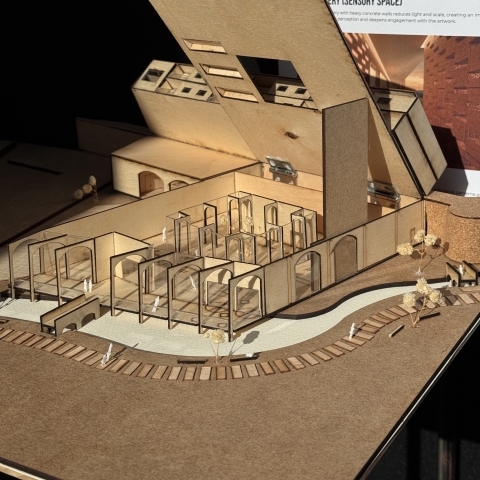 A wooden architectural model with angular forms, an opening slanted roof, and small figures, shown in front of a poster labeled “Sensory Space.”