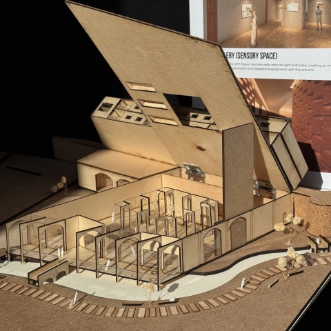 A wooden architectural model with angular forms, an opening slanted roof, and small figures, shown in front of a poster labeled “Sensory Space.”
