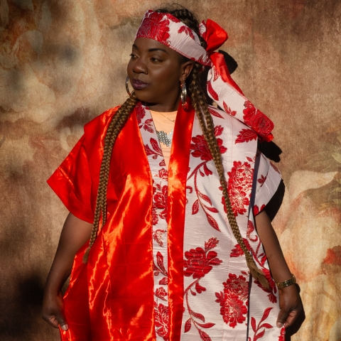 A person stands against a textured brown backdrop wearing a vibrant red and white traditional outfit with floral patterns, along with a matching patterned headwrap topped with a large red bow.