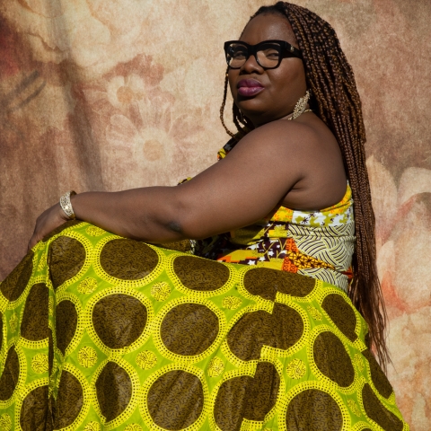 A person wearing vibrant modern African cultural clothing sits sideways against a floral backdrop, with long braids and glasses, reflecting individuality and cultural identity.