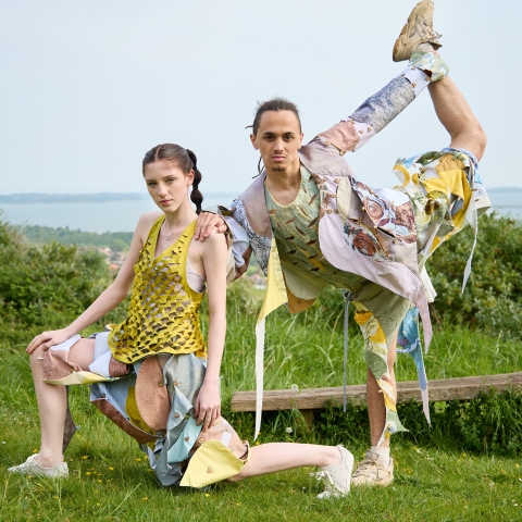 two models posing in a grassy area - one student is lunging forwards the other is holding their leg up behind them - they are wearing garments by Hannah Hatchard