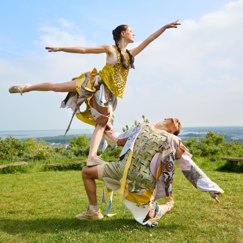 two models in colourful garments in a grassy setting. one setting poses standing on the others knee.