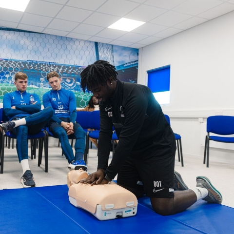 Man giving CPR to dummy in front of seated crowd