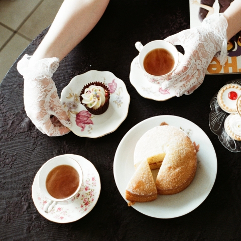 A tea table with two teacups, a small teapot, a sponge cake, jam tarts, and a cupcake. A person wearing white lace gloves holds a teacup and reaches toward the cupcake, with a magazine visible in the corner.