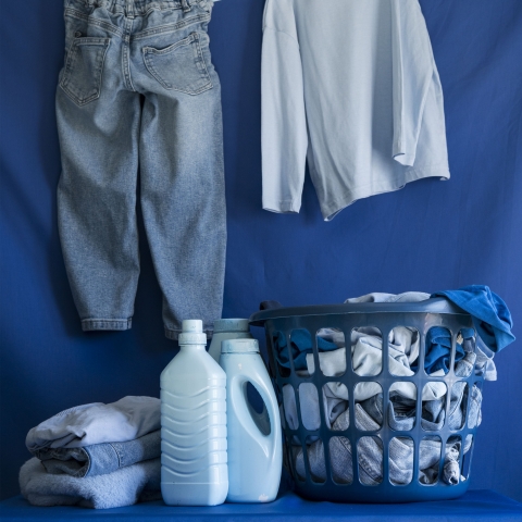 A pair of blue clothes hangs on a line above a laundry basket filled with folded garments, alongside bottles of detergent and towels, forming a monochromatic scene that highlights domestic routines and everyday materials.