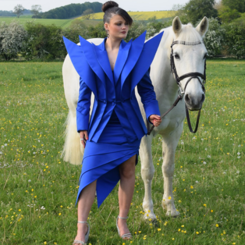 Model wearing a blue garment with pointy shoulder pads - holding a horse in a grassy area