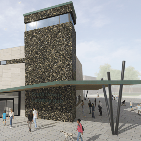 A modern stone‑and‑concrete building with a tall tower, a green canopy entrance, and people walking across the courtyard in front of the South Downs Gateway Hub.