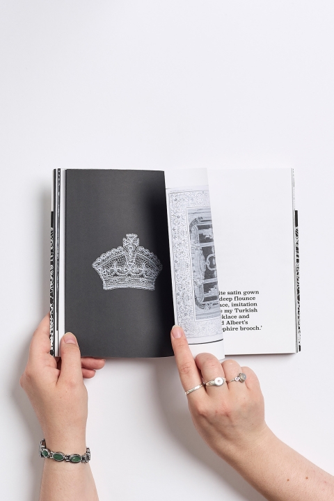 A pair of hands holding an open book, showing an illustrated crown on one page while the other page is being turned to reveal text and an image beneath.