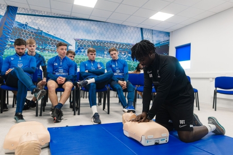 Man giving CPR to dummy in front of seated crowd