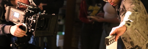 Image of a student behind a camera pointing the camera towards a clapperboard 