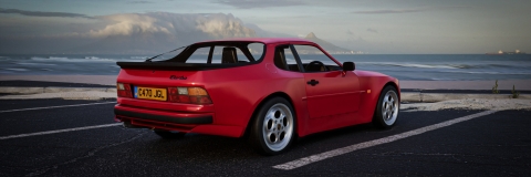 A Computer generated red Porsche win a car park in a snowy setting with the sea in the background. the headlights are open and the car faces towards us