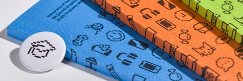 Three brightly coloured hardcover books with pixel‑style icons on the covers, arranged diagonally beside a round pin featuring a pixelated handshake symbol