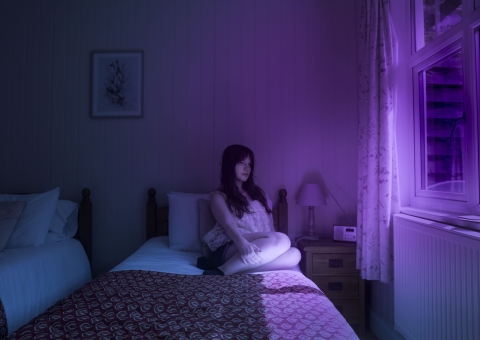A person sits on a bed in a dimly lit wooden‑panelled room, illuminated by vivid purple and blue light from a nearby window. A patterned blanket, a nightstand with a lamp, and a framed picture complete the moody scene.