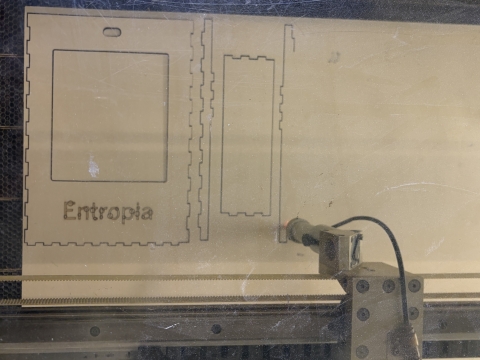 Laser cutter engraving interlocking box components, including a panel with the word “Entropia,” as the machine head moves across the material.