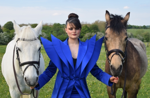 Model with black hair wearing bright blue garment with shoulder pads designed by Isabella Down. The model has a white horse they are holding on the left and a brown horse on the right.