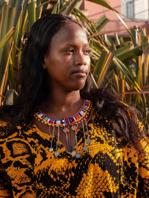 A person stands outdoors in warm sunlight wearing a bright patterned garment and layered beaded jewellery, framed by tall green plants and a pink building, expressing individuality and cultural identity.