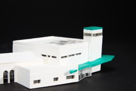 A white architectural model with green roof sections, showing a multi‑level building with a tower, arched openings, and interconnected blocks.