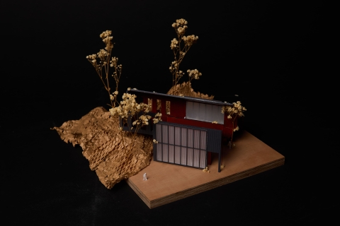 A small architectural model with angled grey and red panels set in textured brown terrain, surrounded by tiny dried plants.