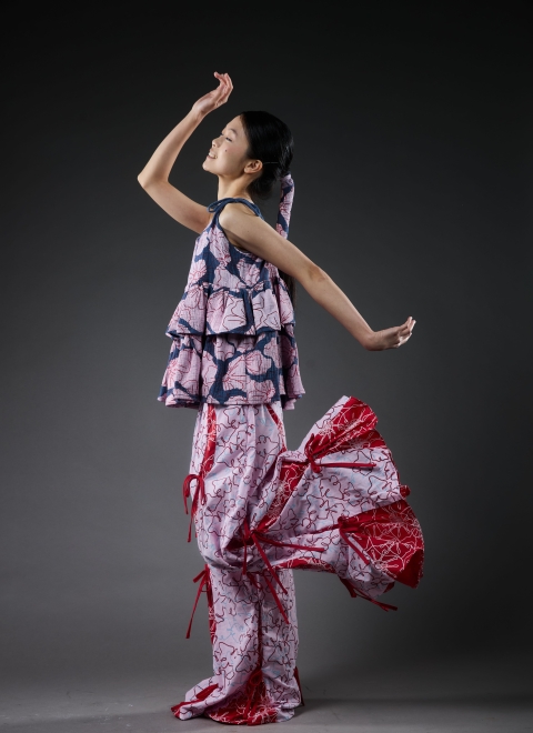 model wearing garment whilst facing left. the garmen is pinky/red at the bottom trousers and the top is more pink/blue. both are quite frilly