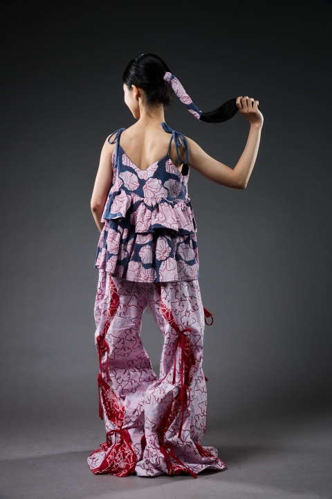 Student wearing garment with pink/red frilly trousers and darker top. there is also a hair piece in the models ponytail in the same material.