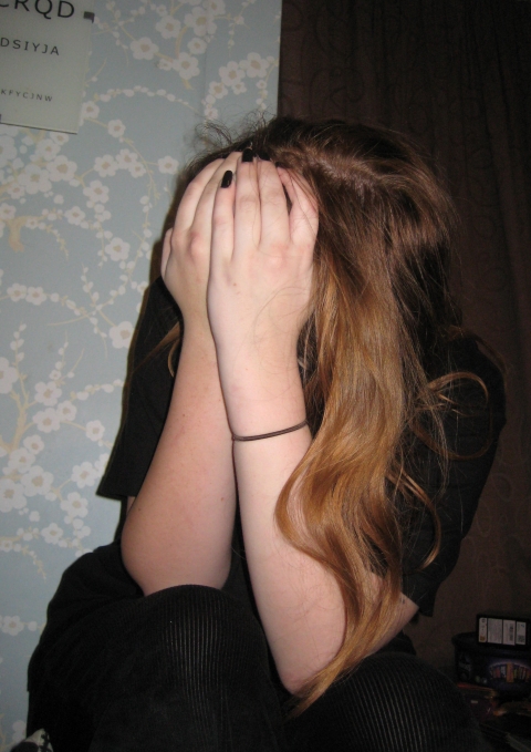 A person sits indoors with long light‑brown hair, covering their face with both hands. A floral patterned wall and a dark curtain form the background.