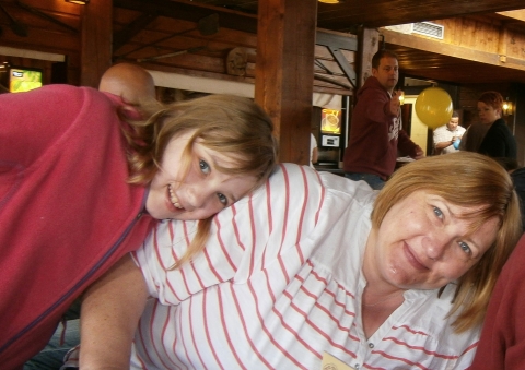 Two people sit close together inside a wooden interior; one leans their head on the other’s shoulder as both smile, with tables, beams and a yellow balloon visible in the background.