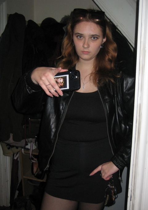 A person takes a mirror selfie indoors, wearing a black leather jacket, black outfit and dark tights, holding a phone with one hand and the other in a pocket. Sunglasses rest on their head, with coats hanging in the background.
