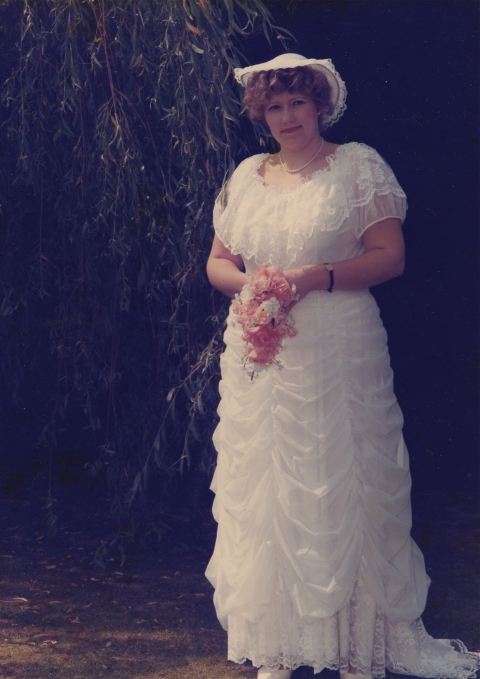 A person stands outdoors wearing a white ruffled gown and holding a small bouquet of pink flowers, with a white headpiece and greenery behind them.