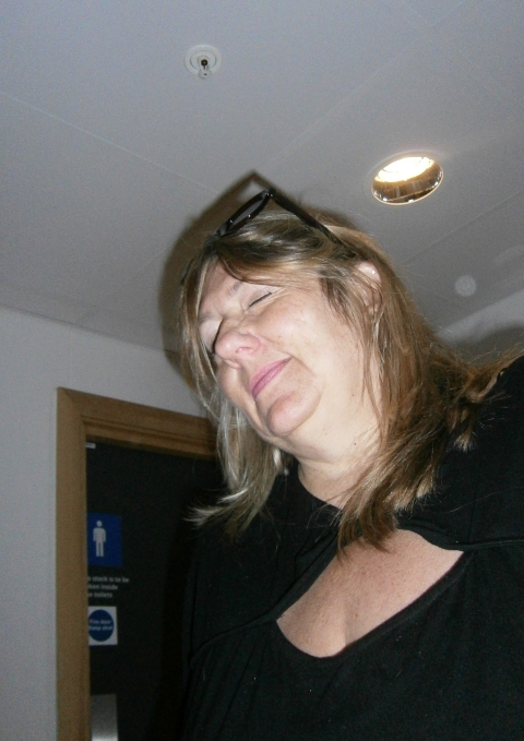A person with long hair stands indoors wearing glasses on their head and a dark shirt, lit by an overhead ceiling light with a doorway and signage visible behind them.