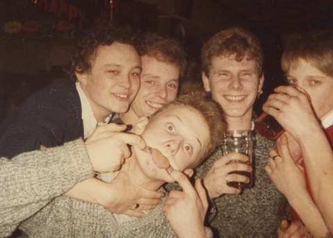 Five people gather closely in a warm indoor setting, some holding drinks, with one person playfully gesturing toward the camera as the group poses together.
