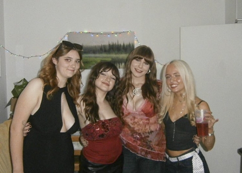 Four people stand close together indoors, dressed for a social occasion, with one holding a drink. A landscape painting and a string of small lights hang on the wall behind them