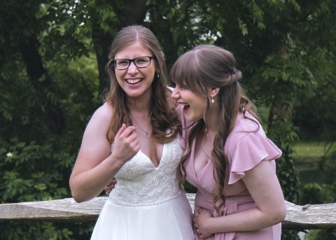 Two people stand outdoors in front of a wooden fence, one in a white lace wedding dress and glasses, the other in a light pink dress, sharing a joyful moment with green trees behind them.