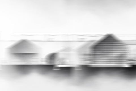 A soft, blurred grayscale scene showing a row of simple house‑like structures with pitched roofs. Their reflections appear faintly below them, creating a misty, dreamlike composition with no sharp details.