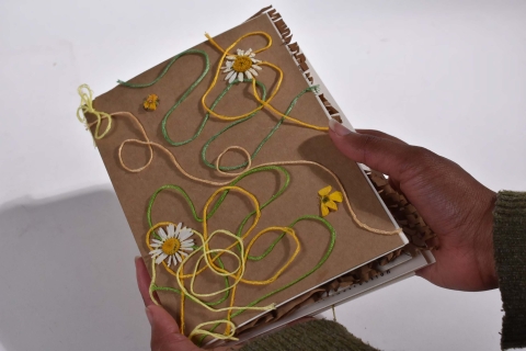 Hands holding a handmade notebook with yarn swirls and small daisy decorations on a brown cover.