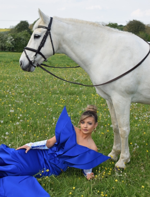 Student laying on grass wearing long blue garment with large shoulder pads - a horse stands above her