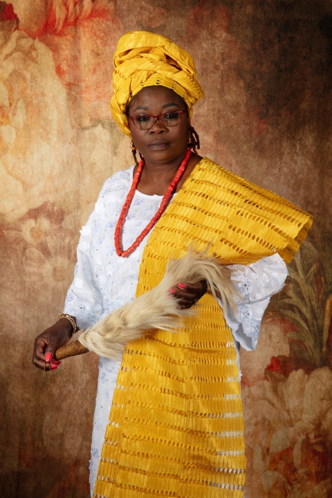 A person wearing elegant West African‑influenced ceremonial clothing stands against a warm floral backdrop, with a bright headwrap, coral‑style beads, and a horsetail whisk, expressing cultural pride and identity.
