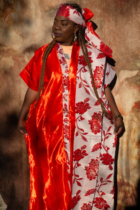 A person wearing bright, patterned ceremonial clothing stands against a textured backdrop, with a matching headwrap, layered fabrics, and jewellery, expressing individuality and cultural identity.