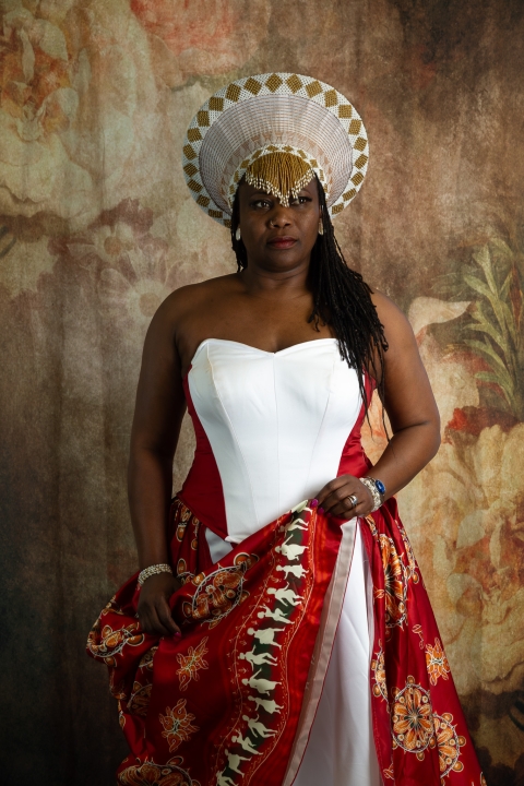 A person wearing ornate ceremonial clothing stands against a floral backdrop, with a large circular headpiece, patterned red‑and‑white garments, long braids, and jewellery, expressing individuality and cultural identity.