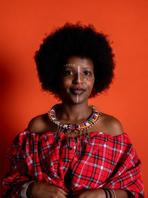 A person stands against a bright orange backdrop wearing a red patterned garment, layered beaded jewellery, white face paint, and an afro hairstyle, expressing individuality and cultural identity.