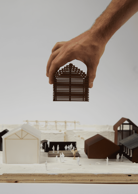 Hand placing a small, arched, brown building model onto a white architectural scale model with several other geometric structures and tiny human figures.
