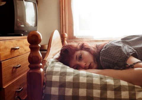 A person lying on a bed with a checkered pillow and blanket, facing the camera. The room has a wooden bed frame, a wooden dresser, and an older TV, lit by sunlight from a window.
