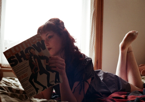A person lying on a bed near a window, reading a magazine titled “SHOW,” resting on their stomach with legs bent upward.