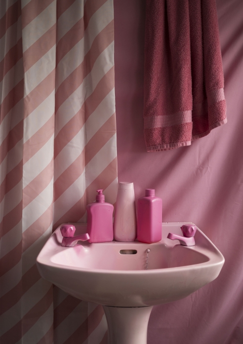 A pink bathroom sink is arranged with matching bottles, towels, and a striped shower curtain, creating a monochromatic scene that highlights the artificial softness and uniformity of domestic products.